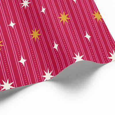 A close-up of a holiday star wrapping paper sheet with a bright pink background, vertical white stripes, and scattered white and yellow starburst motifs, shown on a white background.