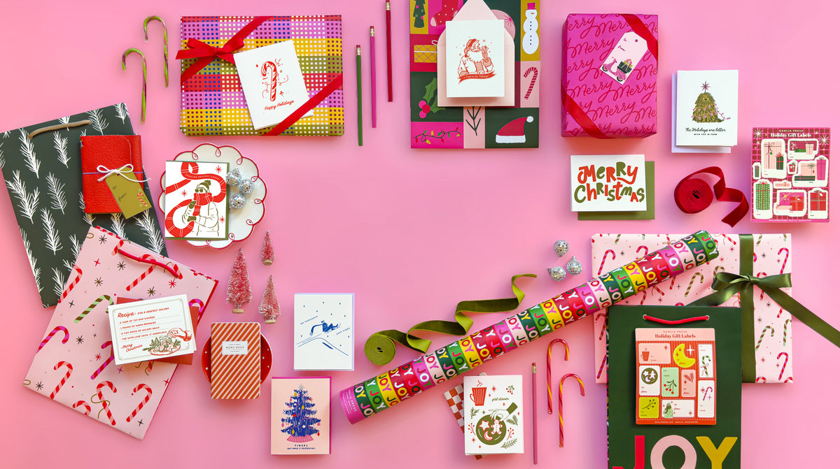 A flat-lay arrangement of various Christmas-themed gift wraps, cards, candy canes, and ribbons on a solid pink background.